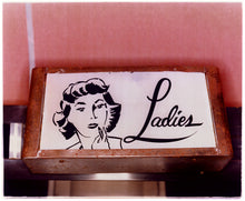 Load image into Gallery viewer, Photograph by Richard Heeps. A kitsch Ladies' toilet sign. The sign has the word Ladies alongside an outline of 1950s woman. The sign sits in a wooden frame against a pink tiled wall.