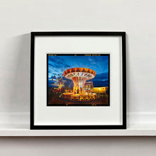 Load image into Gallery viewer, Black framed photograph by Richard Heeps. A fairground ride, the chairoplanes, sits lit in golden and red colours against a dark blue sky.
