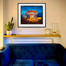 Load image into Gallery viewer, In Situ photograph by Richard Heeps. A fairground ride, the chairoplanes, sits lit in golden and red colours against a dark blue sky.