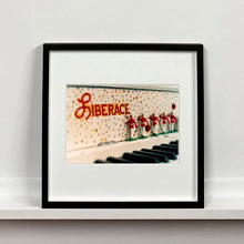 Load image into Gallery viewer, Black framed photograph by Richard Heeps. A close up of Liberace's piano. This captures the black keys and the fall board which is decorated with crystals and red buttons spelling out Liberace in capitals. There are also 5 red and white metal soldiers positioned marching along the rim.