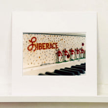 Load image into Gallery viewer, Mounted photograph by Richard Heeps. A close up of Liberace's piano. This captures the black keys and the fall board which is decorated with crystals and red buttons spelling out Liberace in capitals. There are also 5 red and white metal soldiers positioned marching along the rim.