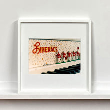 Load image into Gallery viewer, White framed photograph by Richard Heeps. A close up of Liberace's piano. This captures the black keys and the fall board which is decorated with crystals and red buttons spelling out Liberace in capitals. There are also 5 red and white metal soldiers positioned marching along the rim.