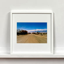 Load image into Gallery viewer, White framed photograph by Richard Heeps. A dusty road in the middle, heading towards the snow capped mountains in the distance, on the right are brown bushes and trees and on the left, single level concrete buildings.