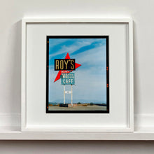 Load image into Gallery viewer, White framed photograph by Richard Heeps edged by film rebate. A roadside sign on Route 66 in America. The word ROY'S appears in a black sign with a bit red arrow pointing to the left ground, below this VACANCY and on a green square the words MOTEL and CAFE.