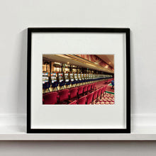Load image into Gallery viewer, Black framed photograph by Richard Heeps. A line of slot machines sits in a vintage Las Vegas casino. Perspective moves the slot machines from big machines on the left hand side to smaller on the right hand side. In front of the machines are a row of neatly lined blue stools one per machine and then red spectators chairs are neatly lined sitting forefront of the shot.