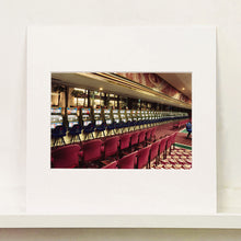 Load image into Gallery viewer, Mounted photograph by Richard Heeps. A line of slot machines sits in a vintage Las Vegas casino. Perspective moves the slot machines from big machines on the left hand side to smaller on the right hand side. In front of the machines are a row of neatly lined blue stools one per machine and then red spectators chairs are neatly lined sitting forefront of the shot.