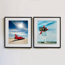 Load image into Gallery viewer, Two black framed photographs by Richard Heeps. The left hand side photograph is of a red drag car with a 75 written on its fin sits on a salt plain the front facing away towards the right. A blue cloudy sky is overhead. The photograph on the right hand side is a large sign for Roy's Motel and Cafe, on flat land with mountains in the background and a vast sky behind.. 