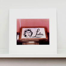 Load image into Gallery viewer, Mounted photograph by Richard Heeps. A kitsch Ladies' toilet sign. The sign has the word Ladies alongside an outline of 1950s woman. The sign sits in a wooden frame against a pink tiled wall.