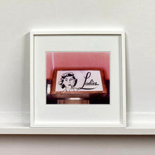 Load image into Gallery viewer, White framed photograph by Richard Heeps. A kitsch Ladies' toilet sign. The sign has the word Ladies alongside an outline of 1950s woman. The sign sits in a wooden frame against a pink tiled wall.