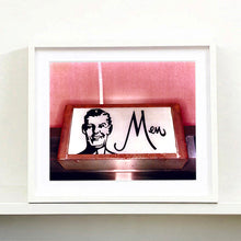 Load image into Gallery viewer, A white framed photograph by Richard Heeps. A kitsch Men's toilet sign. The sign has the word Men alongside an outline of 1950s man. The sign sits in a wooden frame and sits against a pink tiled wall.