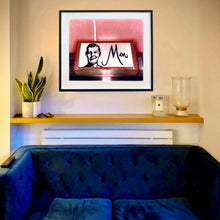 Load image into Gallery viewer, A black framed photograph by Richard Heeps is in situ behind a sofa. The photograph depicts a kitsch Men's toilet sign. The sign has the word Men alongside an outline of 1950s man. The sign sits in a wooden frame and sits against a pink tiled wall.
