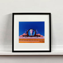 Load image into Gallery viewer, Black framed photograph by Richard Heeps. A 3D shape milkshake parlour sign which has a pink milkshake with a white top, cherry and straws, surrounded by a wooden type shield, and on either side the look of draped American flags. This is set against a blue sky.