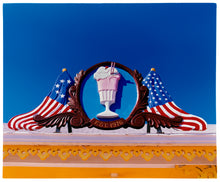 Load image into Gallery viewer, Photograph by Richard Heeps. A 3D shape milkshake parlour sign which has a pink milkshake with a white top, cherry and straws, surrounded by a wooden type shield, and on either side the look of draped American flags. This is set against a blue sky.