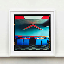 Load image into Gallery viewer, White framed photograph by Richard Heeps. This photograph captures the inside of a Wimpy Restaurant in Norfolk. There is bright blue seats and red tables. The walls are blue and there is a big red chevron light attached to the wall. 