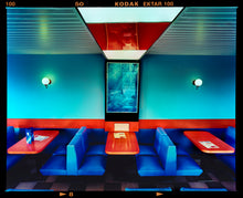 Load image into Gallery viewer, Photograph by Richard Heeps. The inside of a Wimpy restaurant. Blue upholstered double seats pinned to the floor and separated by striking red tables. The walls are blue with a blue picture of trees with a black surround aligned with the middle table.