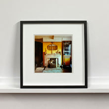 Load image into Gallery viewer, Black framed photograph by Richard Heeps. A vintage room with a fireplace with a brown tiled surround sitting in the middle of the photo flooded in daylight. On the mantelpiece sits 3 candlesticks, the walls behind are painted mustard yellow and there is a a decorative mirror. There is a dresser tucked into the recess on the right hand side.