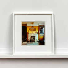 Load image into Gallery viewer, White framed photograph by Richard Heeps. A vintage room with a fireplace with a brown tiled surround sitting in the middle of the photo flooded in daylight. On the mantelpiece sits 3 candlesticks, the walls behind are painted mustard yellow and there is a a decorative mirror. There is a dresser tucked into the recess on the right hand side.