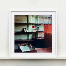Load image into Gallery viewer, White framed photograph by Richard Heeps. The photograph is of an abandoned shoe shop. Four rows of empty shoes shelves curve around a wall, apart from the middle shelf which has two mismatched white stilettos, and a small pile of paraphernalia. The walls are painted light green, held by darker green strutts. Retro brown curtains still hang in the window towards the right of the photograph. 