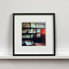 Load image into Gallery viewer, Black framed photograph by Richard Heeps. The photograph is of an abandoned shoe shop. Four rows of empty shoes shelves curve around a wall, apart from the middle shelf which has two mismatched white stilettos, and a small pile of paraphernalia. The walls are painted light green, held by darker green strutts. Retro brown curtains still hang in the window towards the right of the photograph. 