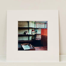 Load image into Gallery viewer, Mounted photograph by Richard Heeps. The photograph is of an abandoned shoe shop. Four rows of empty shoes shelves curve around a wall, apart from the middle shelf which has two mismatched white stilettos, and a small pile of paraphernalia. The walls are painted light green, held by darker green strutts. Retro brown curtains still hang in the window towards the right of the photograph. 