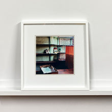 Load image into Gallery viewer, White framed photograph by Richard Heeps. The photograph is of an abandoned shoe shop. Four rows of empty shoes shelves curve around a wall, apart from the middle shelf which has two mismatched white stilettos, and a small pile of paraphernalia. The walls are painted light green, held by darker green strutts. Retro brown curtains still hang in the window towards the right of the photograph. 