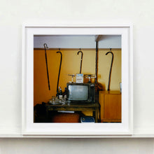 Load image into Gallery viewer, White framed photograph by Richard Heeps. Photograph of a vintage room, a television is on a table with two aerials on top, beside the television are drinks and glasses. Hanging from the picture rail on the yellow wall behind the television are 4 different walking sticks and a bed pan. 