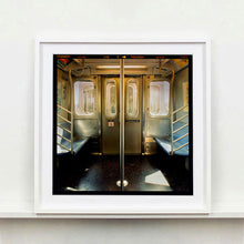 Load image into Gallery viewer, White framed photograph by Richard Heeps. Photograph of the inside of a subway car, looking towards the coaches through the emergency adjoining door. A grab pole sits in the middle.