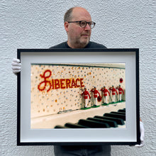 Load image into Gallery viewer, Black framed photograph held by the photographer, Richard Heeps. A close up of Liberace's piano. This captures the black keys and the fall board which is decorated with crystals and red buttons spelling out Liberace in capitals. There are also 5 red and white metal soldiers positioned marching along the rim.