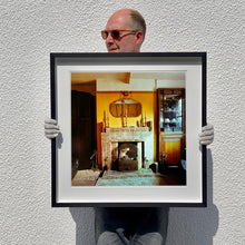 Load image into Gallery viewer, Photograph held by photographer Richard Heeps. A vintage room with a fireplace with a brown tiled surround sitting in the middle of the photo flooded in daylight. On the mantelpiece sits 3 candlesticks, the walls behind are painted mustard yellow and there is a a decorative mirror. There is a dresser tucked into the recess on the right hand side.