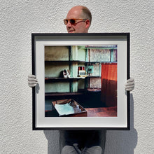Load image into Gallery viewer, Black framed photograph held by photographer Richard Heeps. The photograph is of an abandoned shoe shop. Four rows of empty shoes shelves curve around a wall, apart from the middle shelf which has two mismatched white stilettos, and a small pile of paraphernalia. The walls are painted light green, held by darker green strutts. Retro brown curtains still hang in the window towards the right of the photograph. 