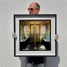 Load image into Gallery viewer, Black framed photograph held by the photographer Richard Heeps. Photograph of the inside of a subway car, looking towards the coaches through the emergency adjoining door. A grab pole sits in the middle.