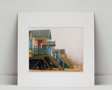 Load image into Gallery viewer, 'Beach Huts', photographed by Richard Heeps in one of his favourite areas of the UK, the port town of Wells-next-the-sea, Norfolk. This piece captures the classic quintessential beach hut on an atmospherically foggy day.