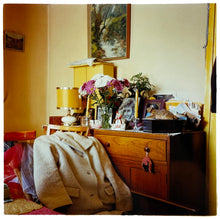 Load image into Gallery viewer, Photograph by Richard Heeps. A wooden dresser with photos, bunches of flowers, candles, ornaments, lamps and a full inbox on top.  In front of this busy piece of furniture are clothes laid on a chair, on top a white fur jacket. 