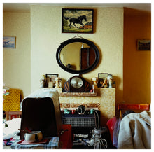 Load image into Gallery viewer, Photograph by Richard Heeps. A mantelpiece is in the middle of this photograph with an electric fire sitting in its hearth.  The mantelpiece is surrounded and covered by paraphernalia. Above the mantelpiece is a mirror and above that a picture of a black horse. There is a boxed sewing machine in front of the mantelpiece.  