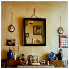 Load image into Gallery viewer, Photograph by Richard Heeps. A tiled mantelpiece full of paraphernalia from the past: clocks; porcelain animals and curios all seemingly randomly displayed.  There is a square mirror over the mantelpiece reflecting the room and on the far right of the photo is a red and white calendar, displaying July 1988. 