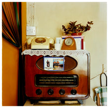 Load image into Gallery viewer, Photograph by Richard Heeps. Daisy's radio in its wooden casing sits in the middle of this photograph.  There is a postcard from Largs stuck on the radio speakers and on top of the radio the surface is crowded with houseplants, a phone, a mantel clock and other oddments. 