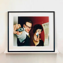 Load image into Gallery viewer, A couple look into the camera in burlesque costume against a red wall. Contemporary portrait photography by Richard Heeps framed in black