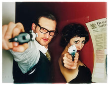 Load image into Gallery viewer, A couple look into the camera in burlesque costume against a red wall. Contemporary portrait photography by Richard Heeps