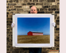 Load image into Gallery viewer, 'Stars &amp; Stripes Barn' shows a depiction of the American flag painted on this isolated building in Oakhurst, California. It sits on the horizon against a vast bright blue sky. This artwork is part of Richard Heeps' 'Dream in Colour' series.