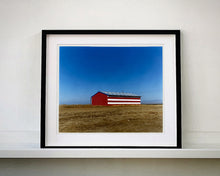 Load image into Gallery viewer, 'Stars &amp; Stripes Barn' shows a depiction of the American flag painted on this isolated building in Oakhurst, California. It sits on the horizon against a vast bright blue sky. This artwork is part of Richard Heeps' 'Dream in Colour' series.