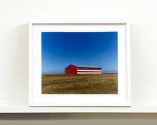 Load image into Gallery viewer, 'Stars &amp; Stripes Barn' shows a depiction of the American flag painted on this isolated building in Oakhurst, California. It sits on the horizon against a vast bright blue sky. This artwork is part of Richard Heeps' 'Dream in Colour' series.