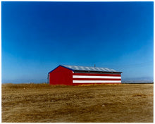 Load image into Gallery viewer, 'Stars &amp; Stripes Barn' shows a depiction of the American flag painted on this isolated building in Oakhurst, California. It sits on the horizon against a vast bright blue sky. This artwork is part of Richard Heeps' 'Dream in Colour' series.